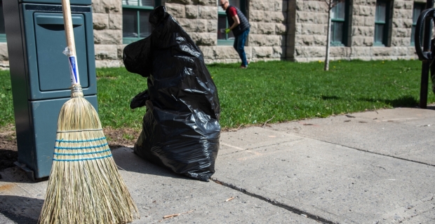 Un balai et un sac-poubelle en plastique à moitié plein sur le trottoir avec un homme ramassant des déchets en arrière-plan