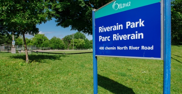 A sign for Riverain Park with a baseball diamond in the background