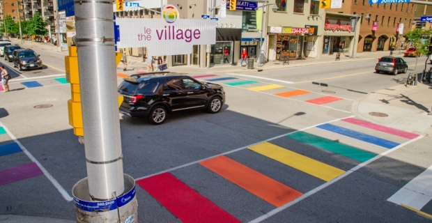 Un passage piéton peint aux couleurs de l’arc-en-ciel au centre-ville avec un panneau indiquant « le village »